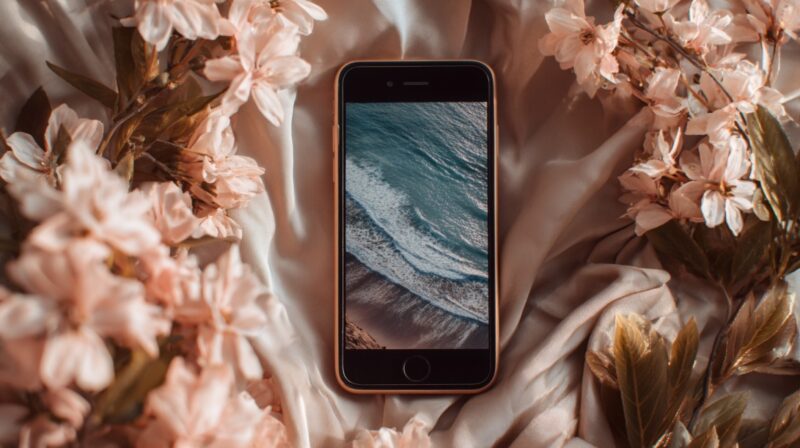 A smartphone displaying an ocean wave on its screen, surrounded by soft pink flowers and satin fabric