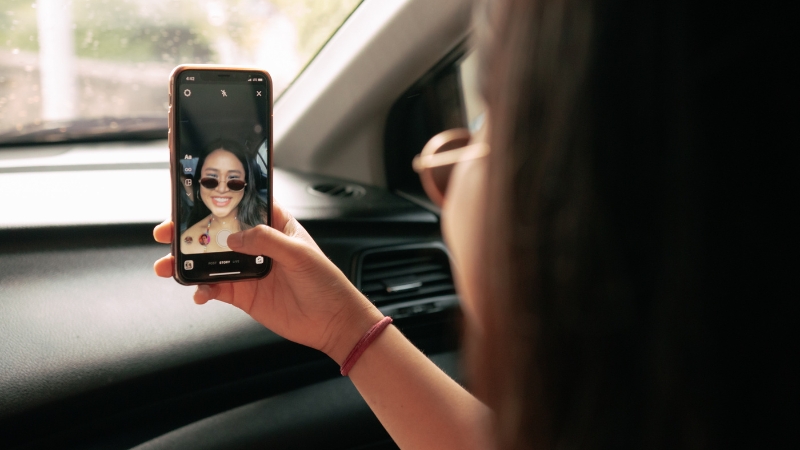 A woman records an Instagram Reel on her phone inside a car, smiling at the camera