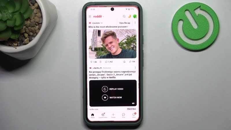 A smartphone displays the Reddit app interface with a post and ad visible, placed on a desk